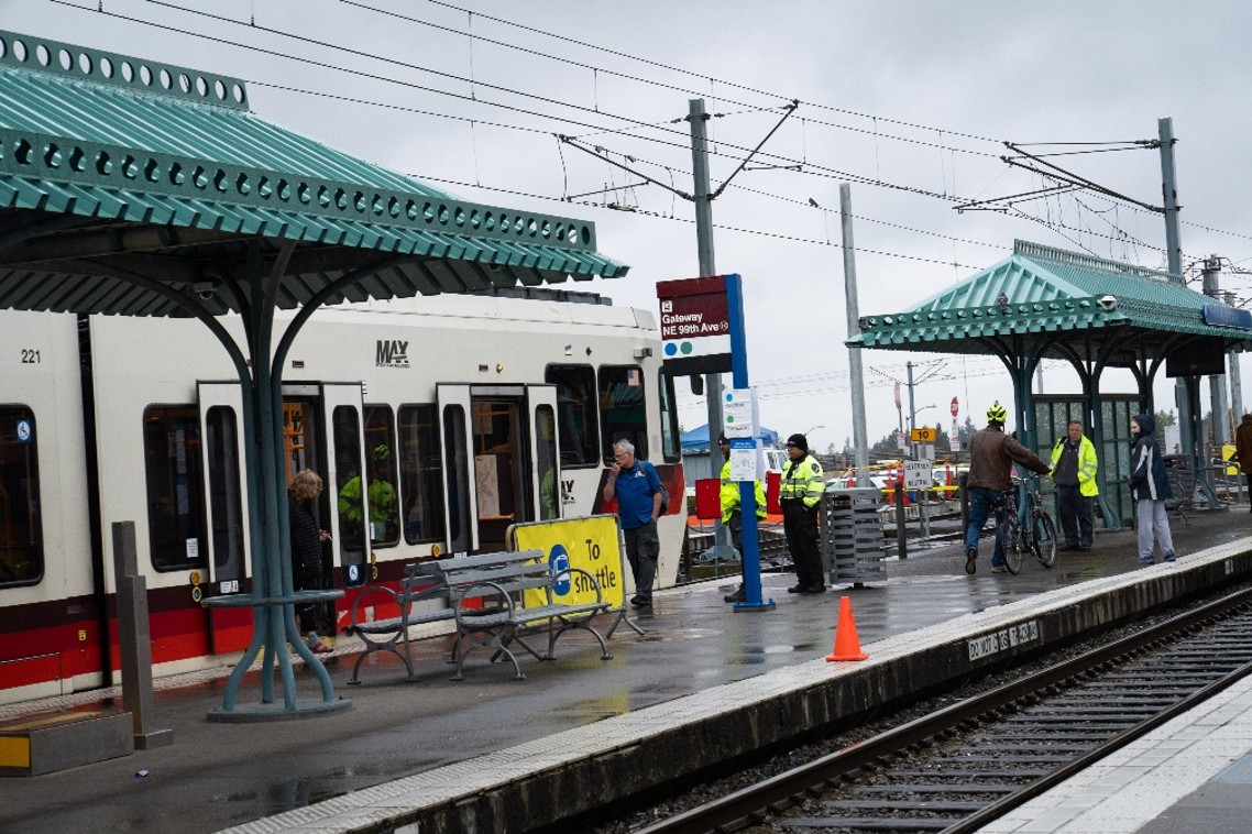 People wearing safety vests assist passengers on a light rail platform during rainy weather. A train is stopped at the station, with benches, platform shelters, and rail tracks visible in the foreground. Photo credit: Ride Connection, Portland, OR.